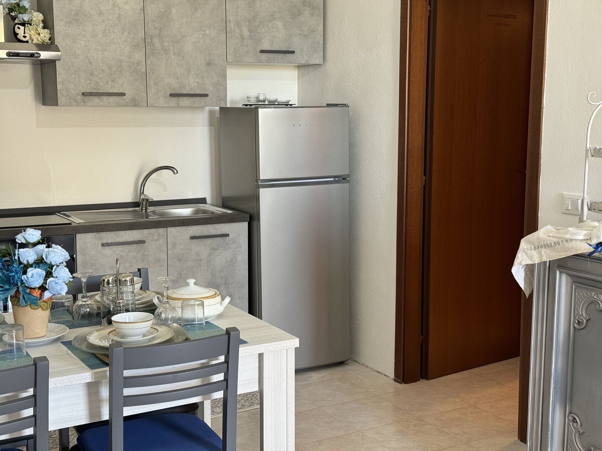 Een kleine keuken met een koelkast, gootsteen en kastjes. Er staat een eettafel met stoelen en een bloemenvaas.