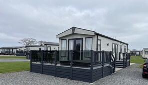 A mobile home with a deck and a covered porch, surrounded by a gravel driveway and a grassy lawn.