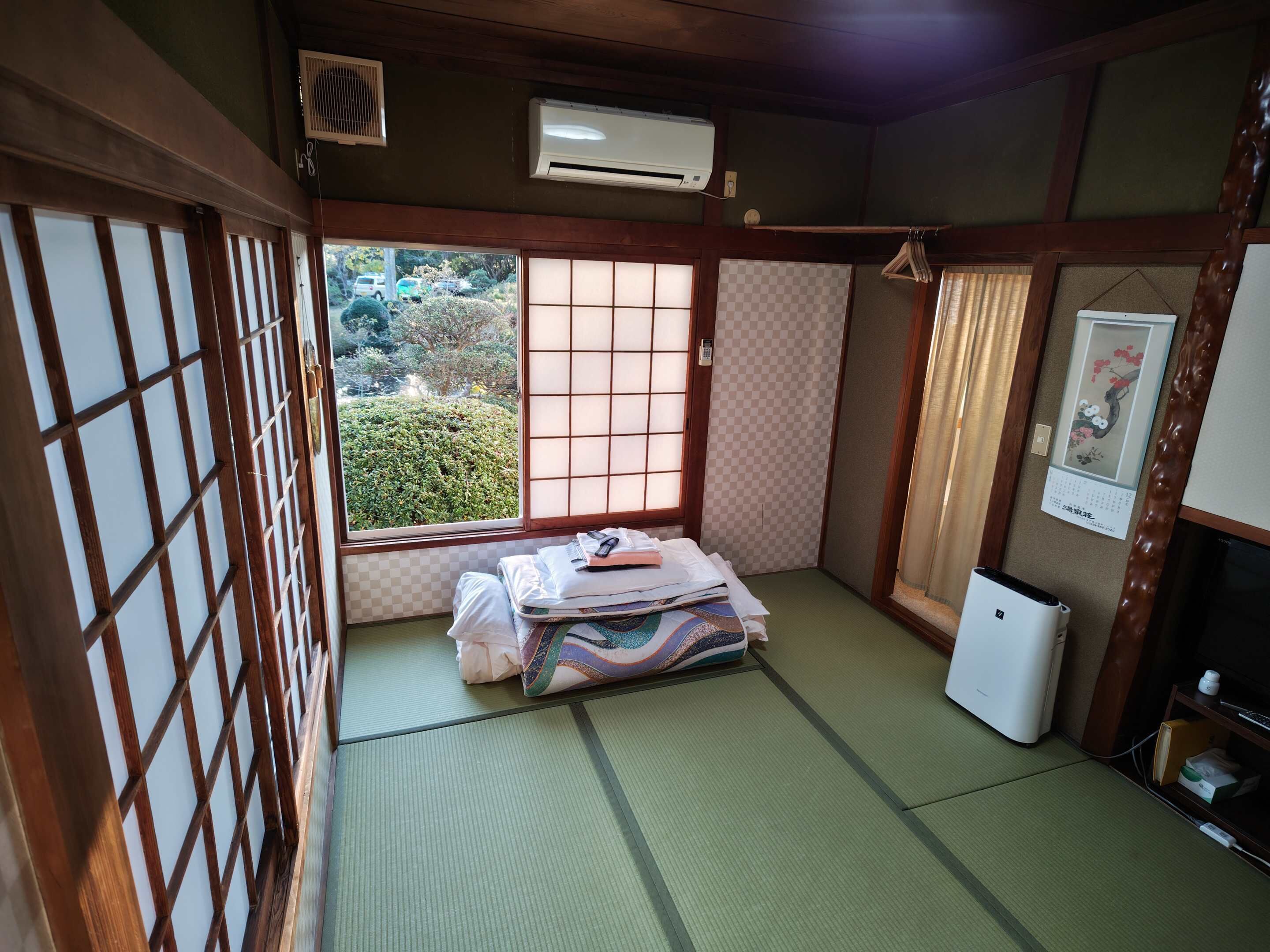 Una tradizionale stanza giapponese con pavimento in tatami, un futon e un piccolo condizionatore d'aria, con vista su piante verdi.