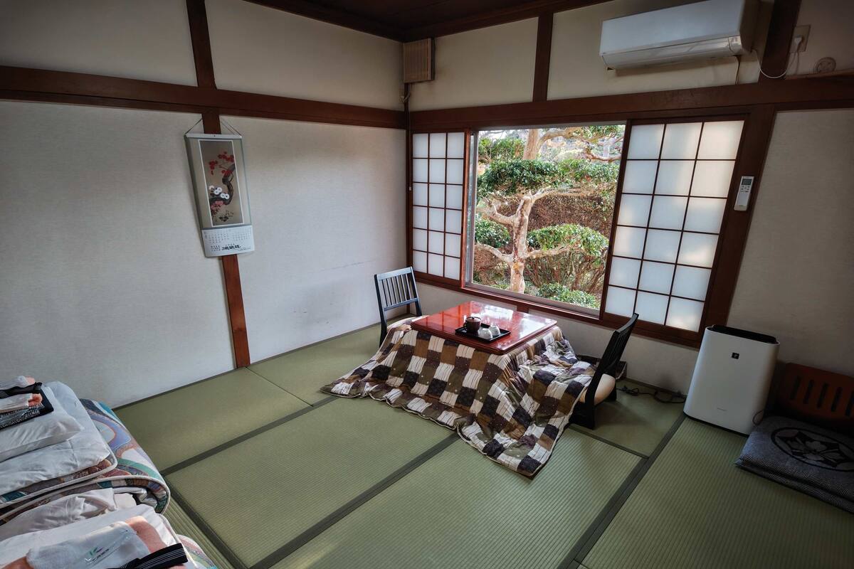 A traditional Japanese room with tatami flooring, a low table with a cushion, a chair, and a view of a garden through shoji screens.