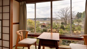 Ein Zimmer mit Tatami-Boden, einem Holztisch, zwei Stühlen und einem großen Fenster mit Blick auf einen Garten und Gebäude.