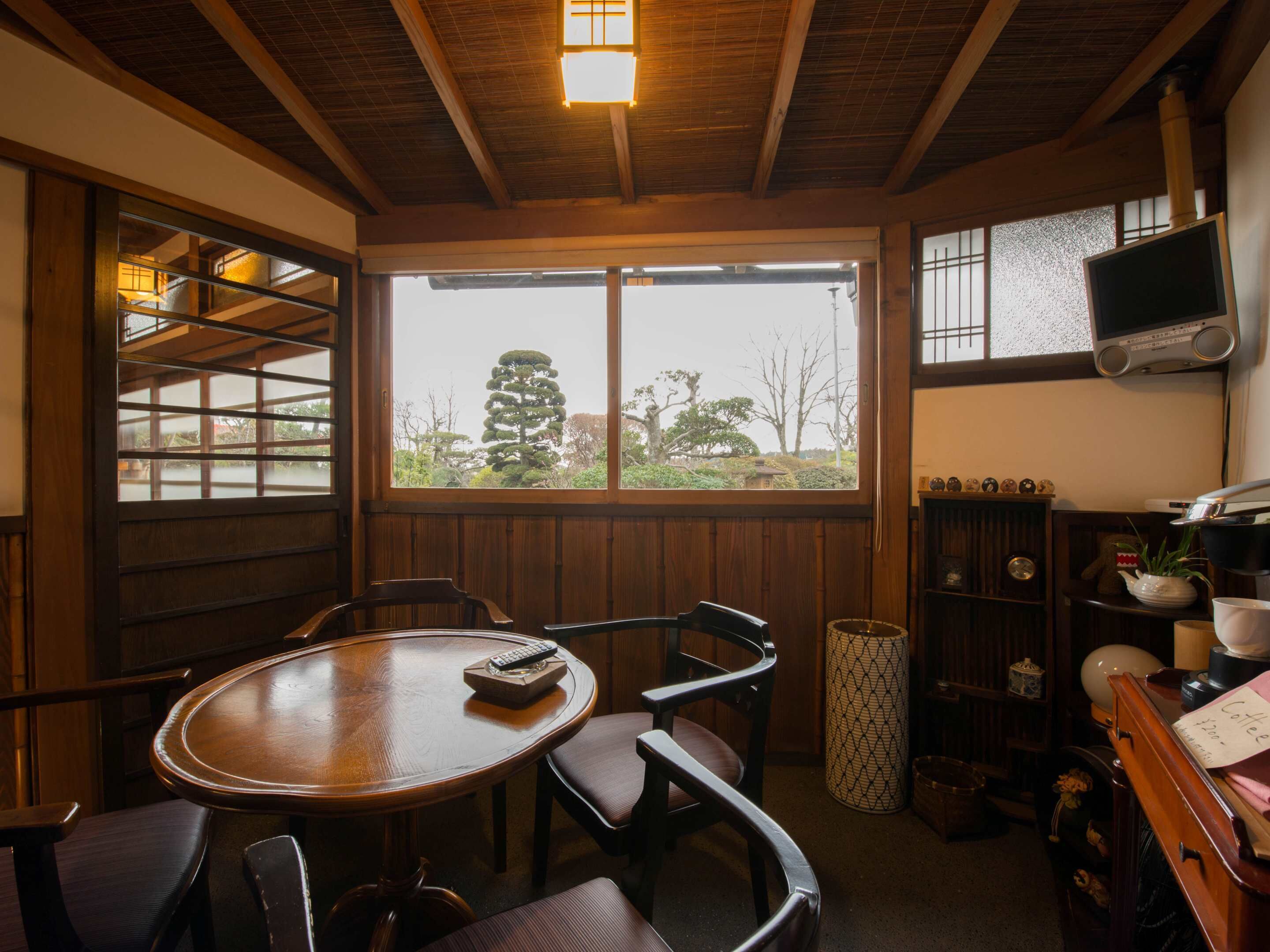 A cozy room with a wooden ceiling, a round table with chairs, a TV mounted on the wall, and a window with a view of trees.
