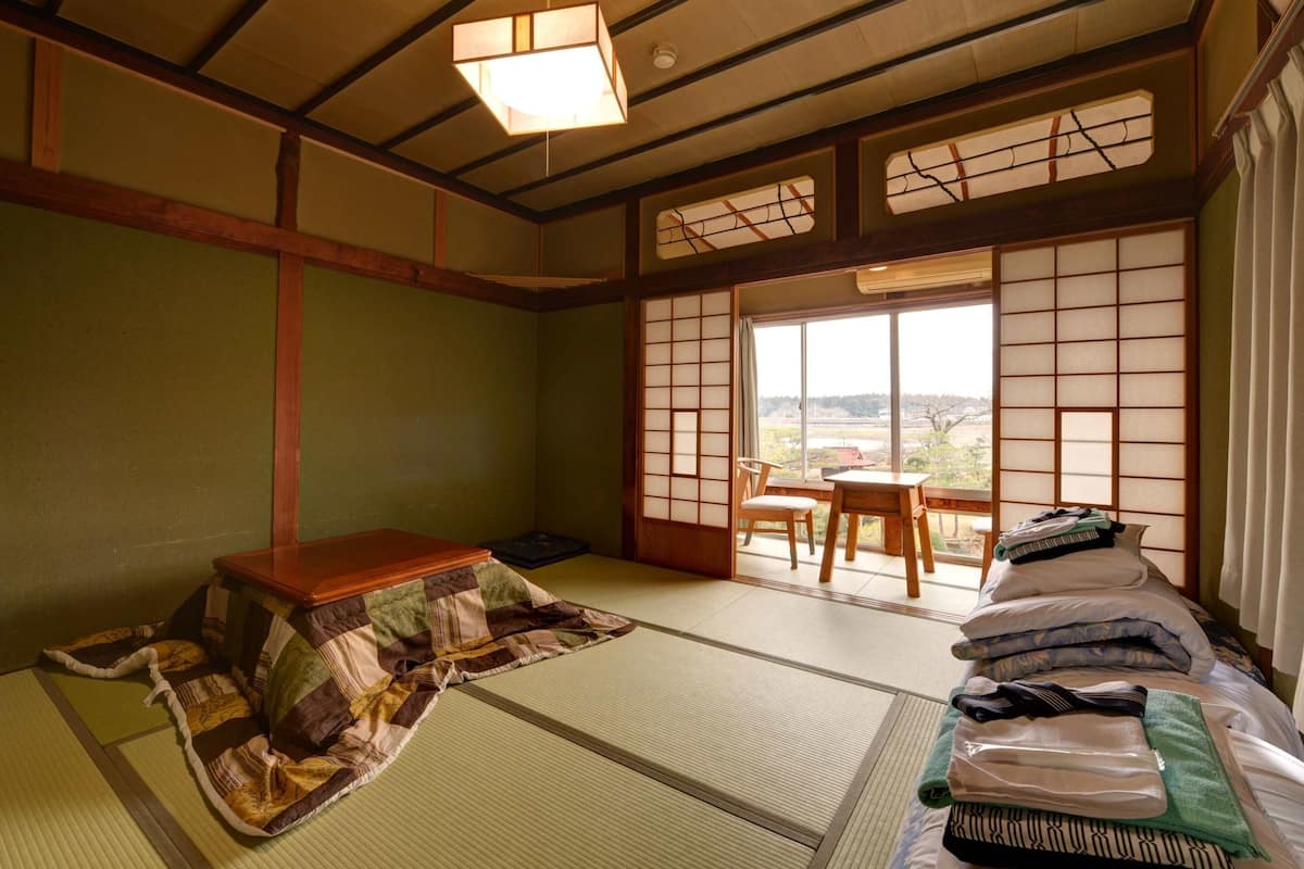 A traditional Japanese room with tatami flooring, a low wooden table, a futon bed, and a sliding shoji screen door leading to a balcony with a view of the countryside.