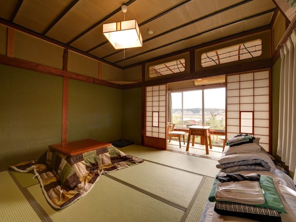 A traditional Japanese room with tatami flooring, a low wooden table, a futon bed, and a sliding shoji screen door leading to a balcony with a view of the countryside.