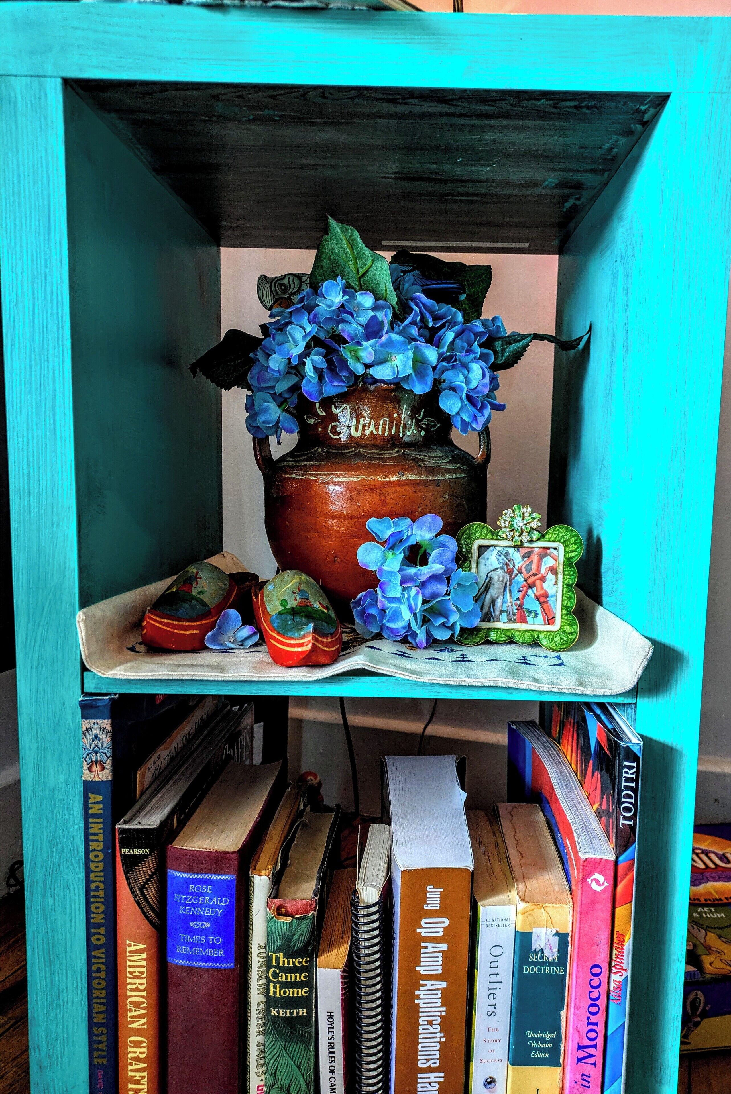 A bookshelf with a variety of books, a ceramic pot with blue flowers, and a framed picture.