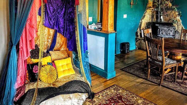 Una habitación con una cama cubierta de tejidos coloridos, una mesa de madera con sillas y una chimenea de piedra.