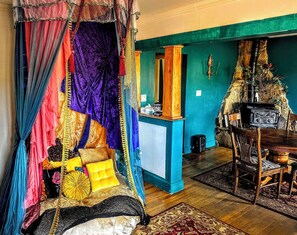 Une pièce avec un lit recouvert de tissus aux couleurs vives, une table en bois avec des chaises et une cheminée en pierre.