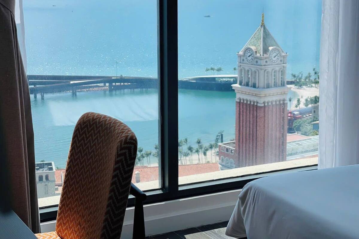 A hotel room with a large window offering a view of a coastal cityscape, including a prominent clock tower and a bridge.
