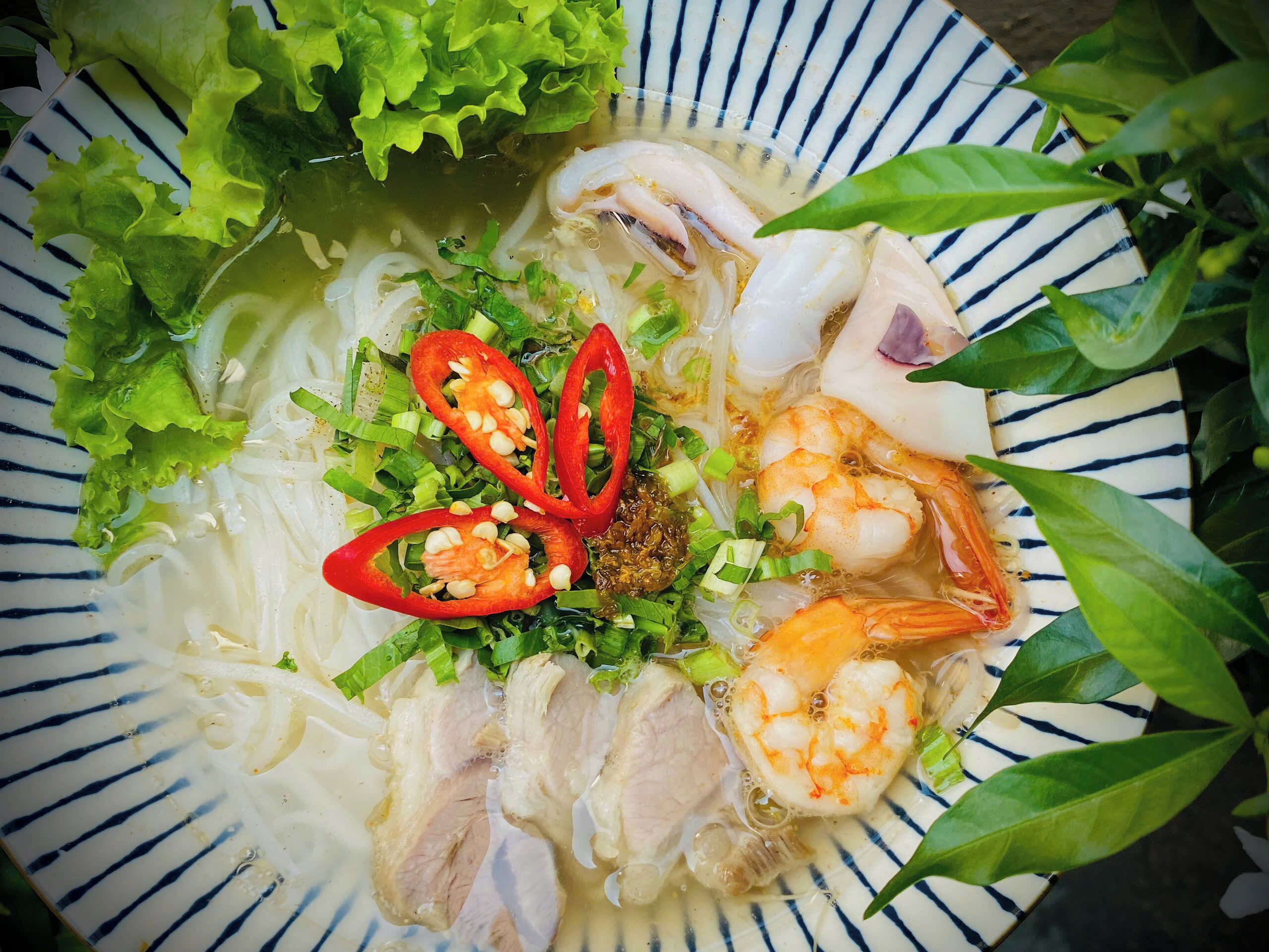 A bowl of noodle soup with shrimp, lettuce, and sliced red chili peppers.