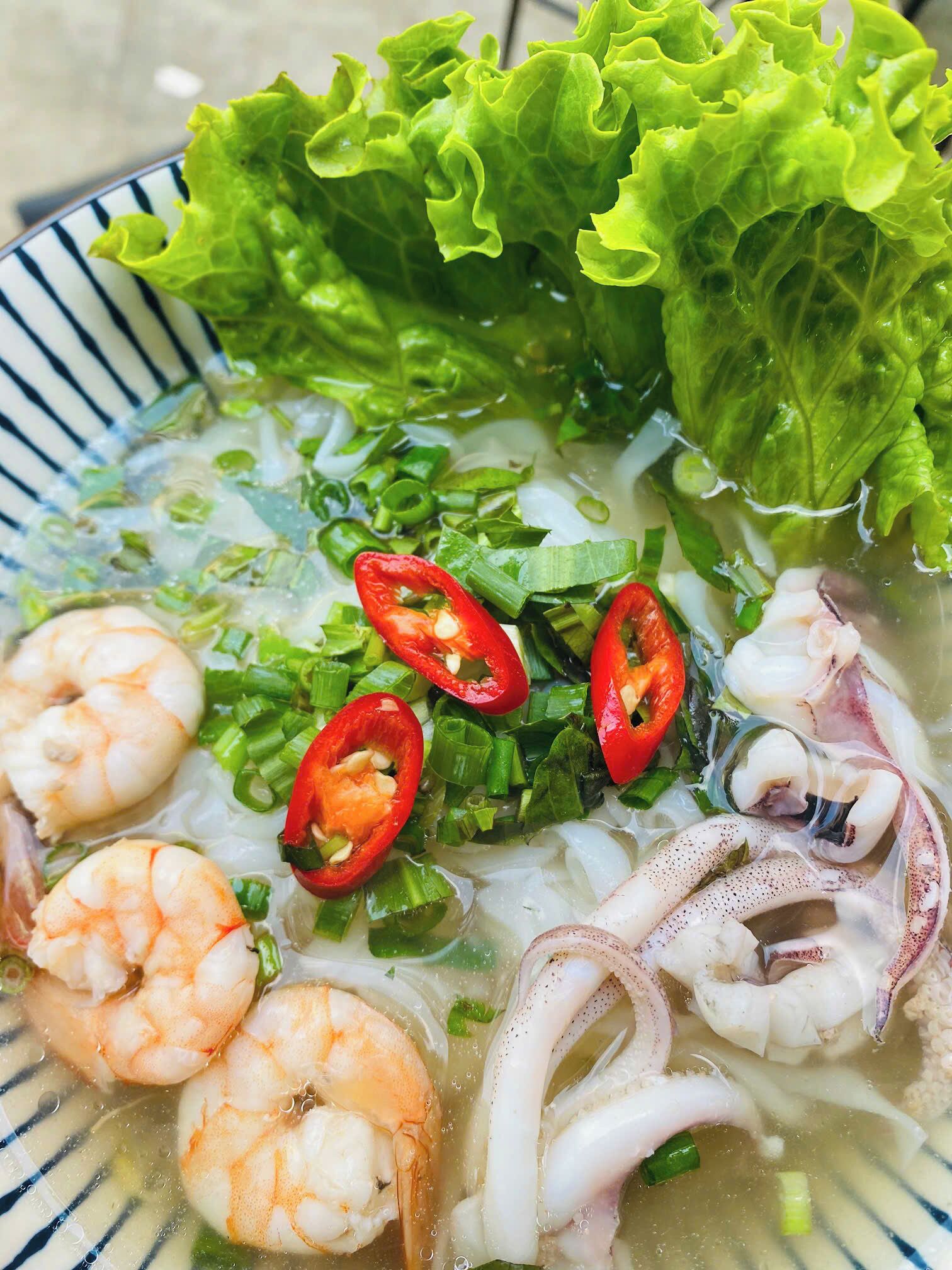 A bowl of seafood soup with shrimp, squid, and octopus, garnished with red chili and green onions.