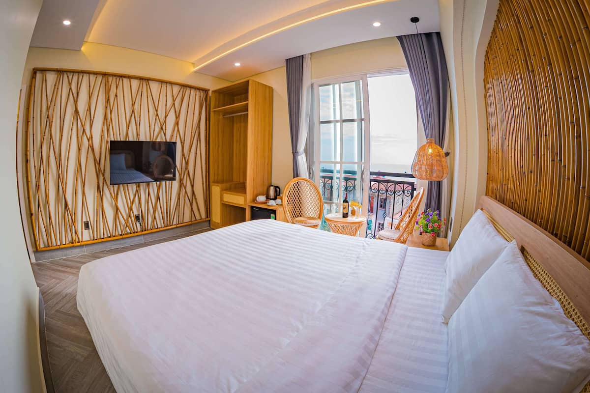 A hotel room with a large bed, a TV mounted on the wall, a balcony with wicker furniture, and a view of the city.