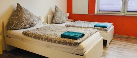 A room with two beds, each with patterned bedding and pillows, a window with blinds, and a red wall.