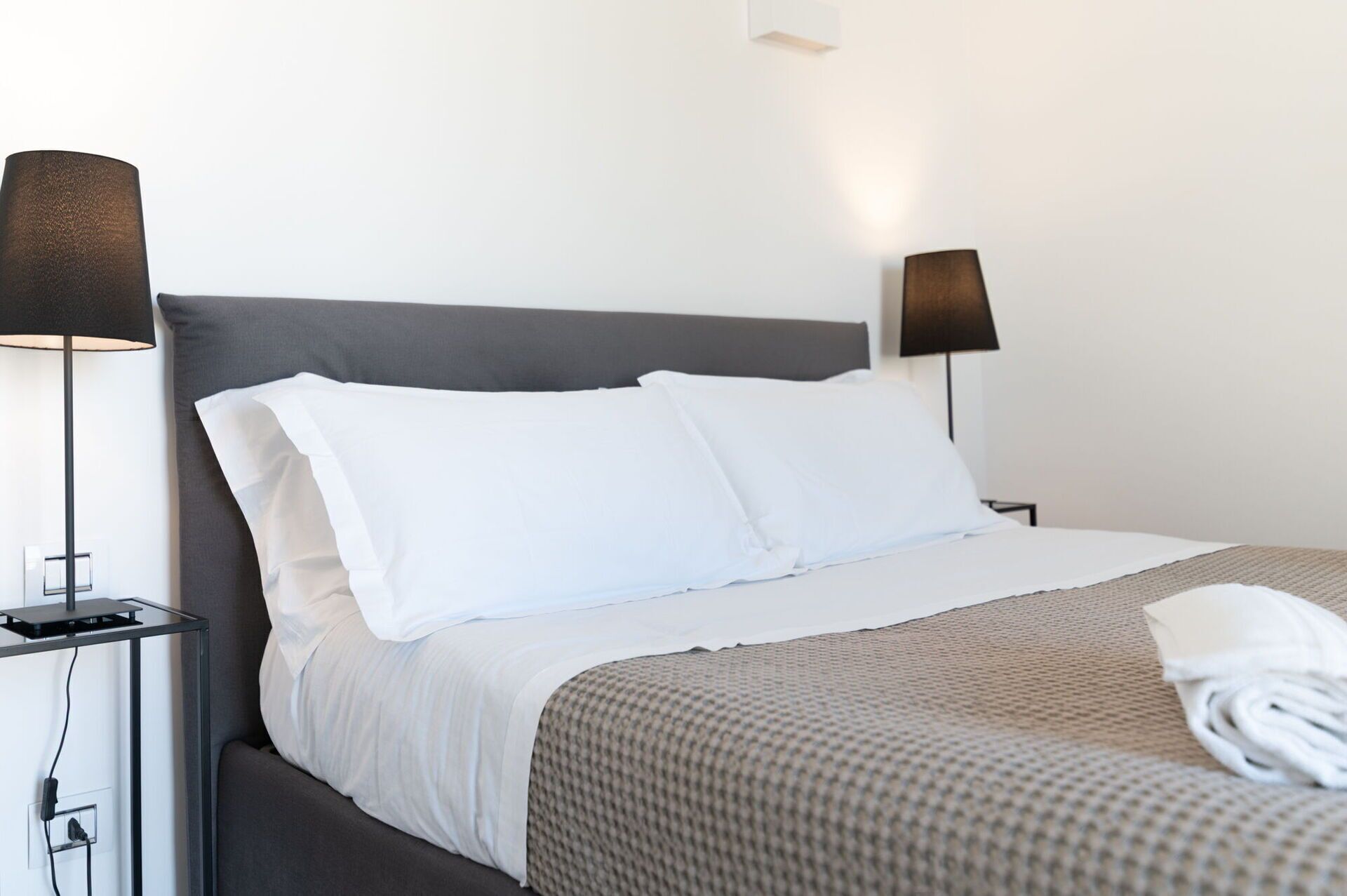 A neatly made bed with white linens and a gray headboard, flanked by two bedside tables with lamps.