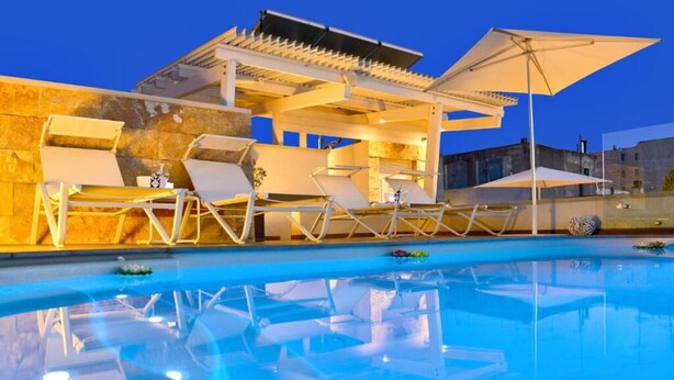 Una piscina con sillas de descanso y sombrillas, rodeada de muros de piedra y un edificio con techo inclinado.