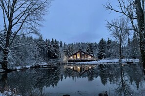 A cabin with a reflection in a calm body of water, surrounded by snow-covered trees.