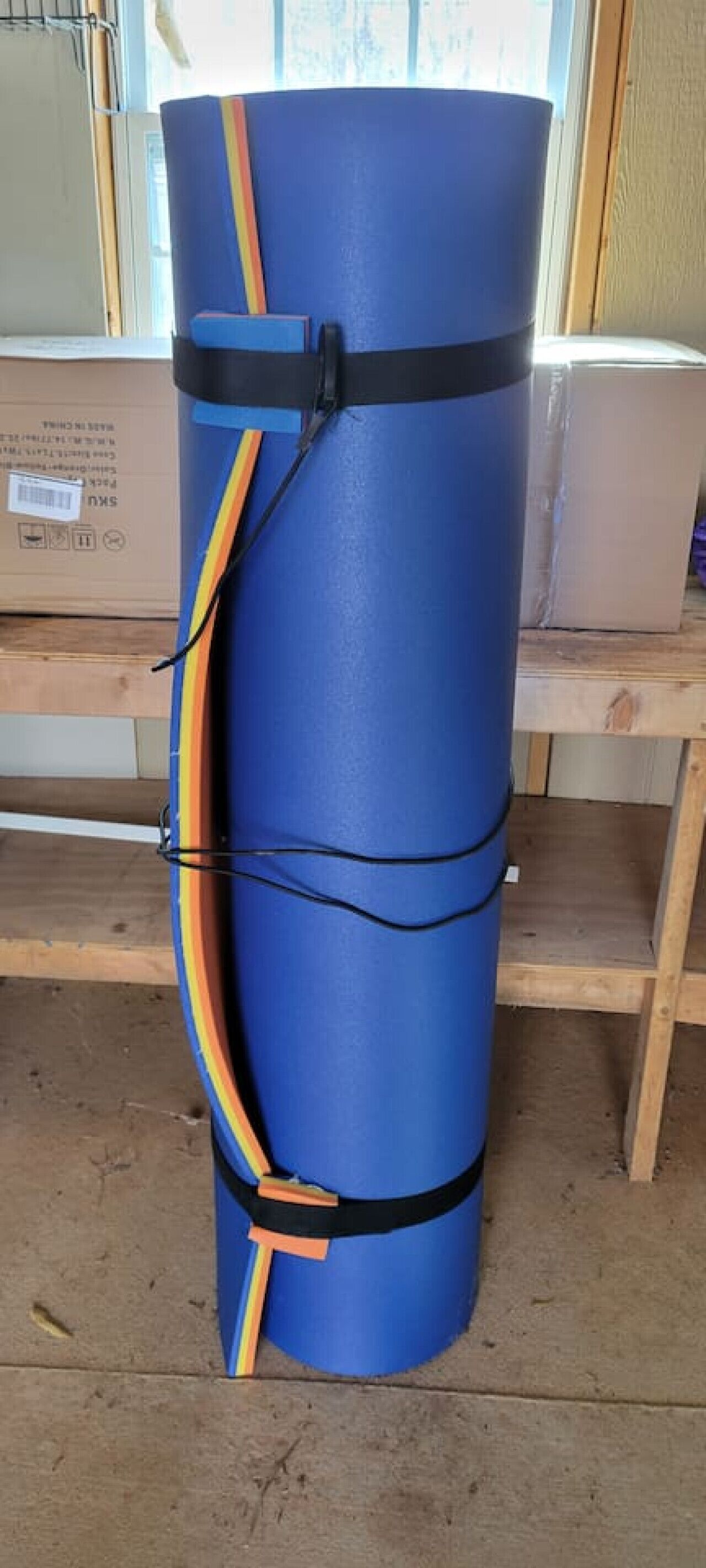 A blue cylindrical object with black and orange straps, placed on a wooden floor in a room with a window and a shelf in the background.