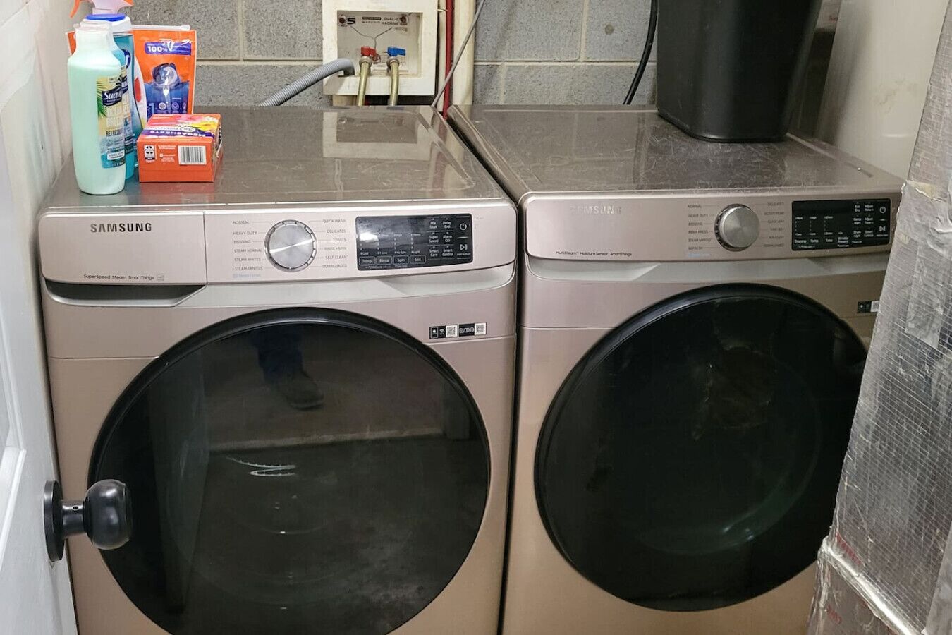 Two Samsung front-loading washers in a laundry room with a tiled wall and a countertop holding cleaning supplies.