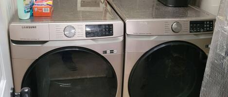 Two Samsung front-loading washers in a laundry room with a tiled wall and a countertop holding cleaning supplies.
