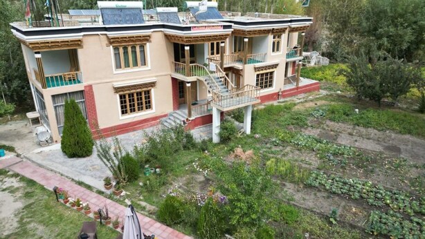 A two-story building with balconies, a central staircase, and a garden with various plants and flowers. - The Latsas (Hunder)