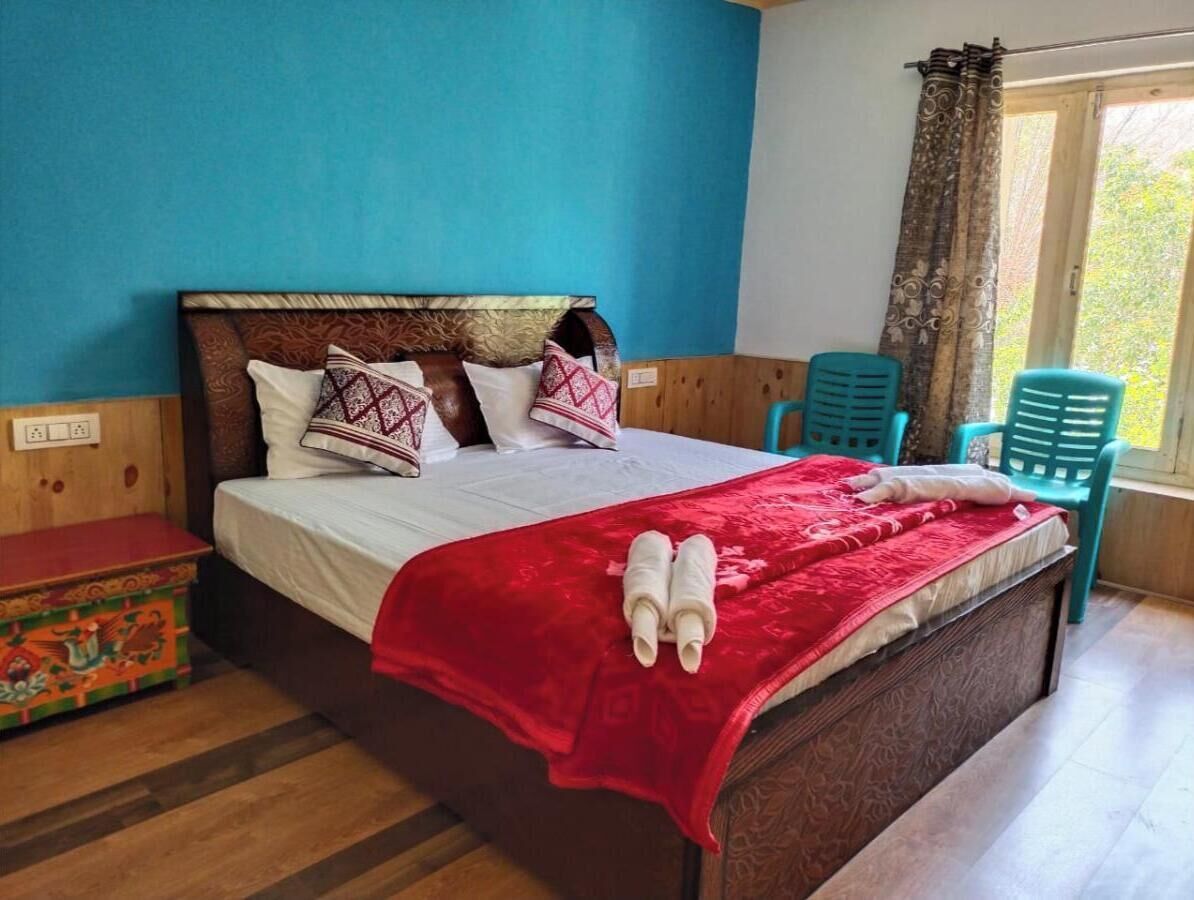 A neatly made bed with a red blanket and two towels, a wooden headboard, and a small wooden chest of drawers.