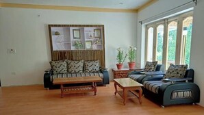 A living room with a sofa, a coffee table, and a set of chairs. There is a wall-mounted shelf with decorative items and a window with a view of greenery. - The Latsas (Hunder)