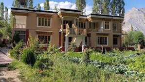 A two-story building with a balcony, surrounded by greenery and mountains in the background. - The Latsas (Hunder)