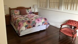 A bedroom with a floral bedspread, a wooden headboard, a nightstand with a lamp, and a red upholstered chair. - Library Cottage (Hamilton)