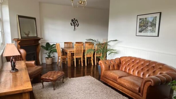 A living room with a brown leather sofa, wooden coffee table, and a fireplace. There are two wooden chairs and a dining table in the background. - Library Cottage (Hamilton)