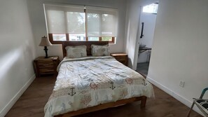 Un dormitorio con cama, mesitas de noche, lámpara, ventana con persianas y vista al exterior.