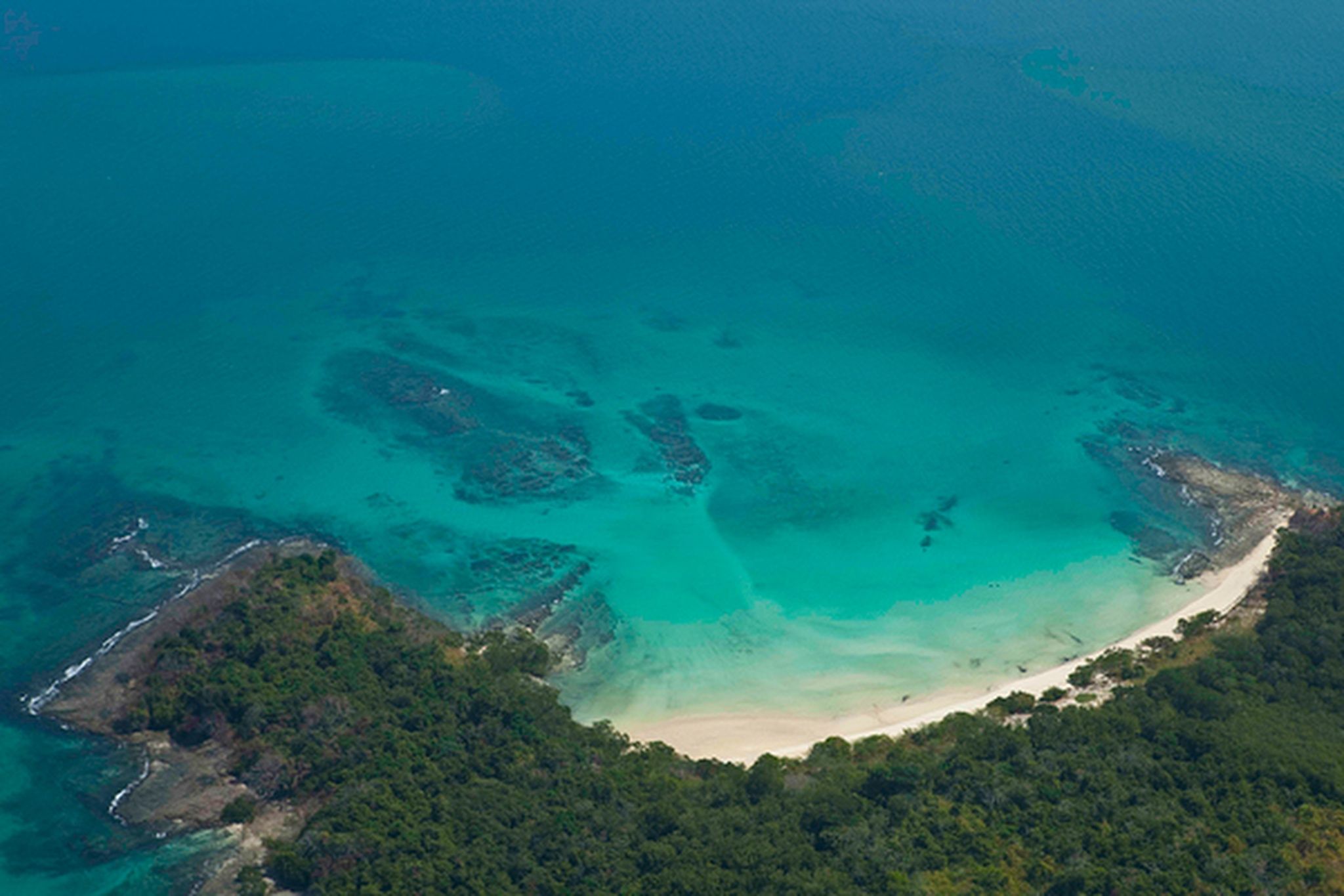 Vista aérea de una zona costera con aguas turquesas cristalinas, una playa de arena y vegetación verde exuberante.