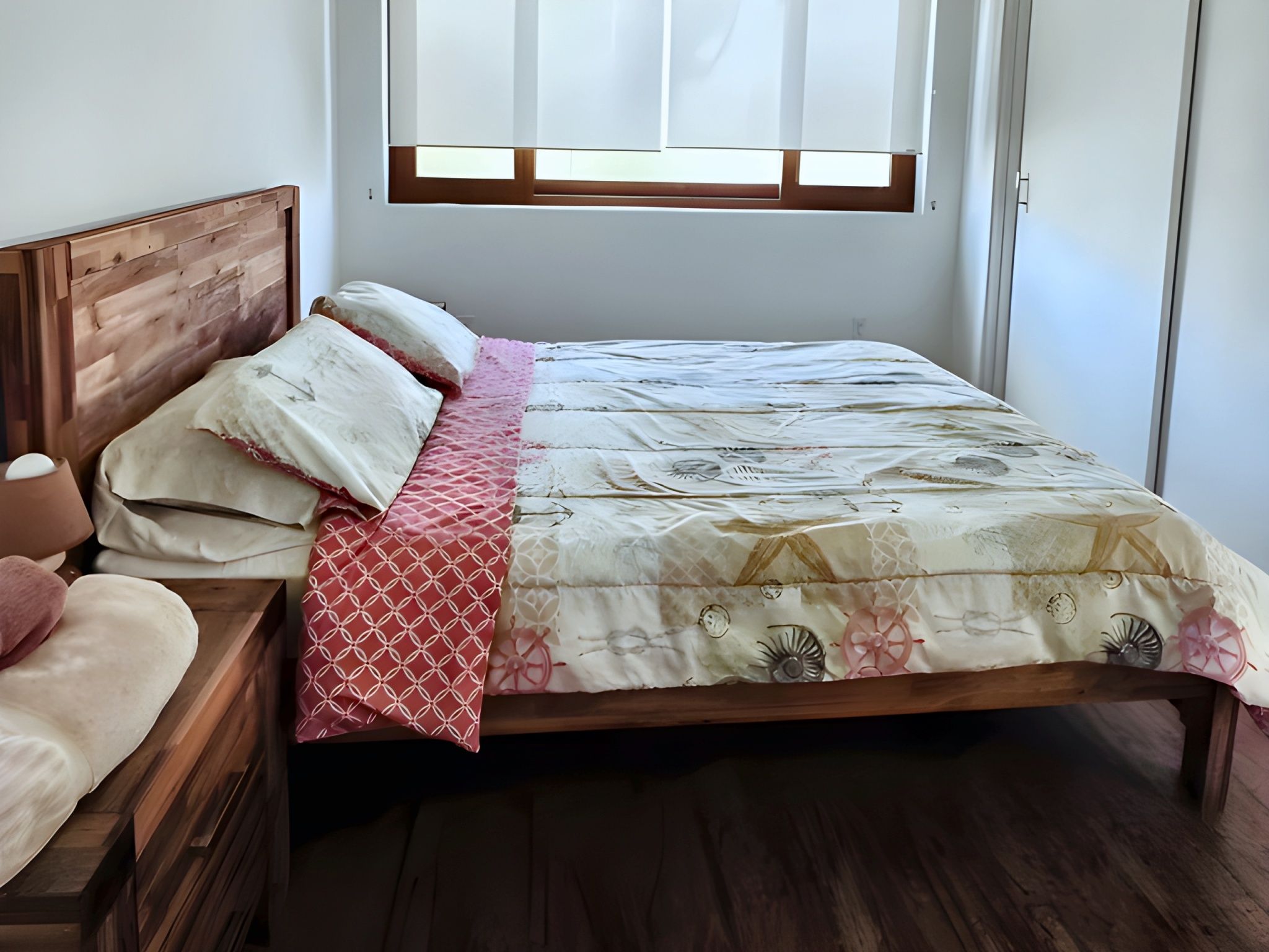 A bedroom with a wooden headboard, a bed with a patterned bedspread, a bedside table with a lamp, and a window with blinds.