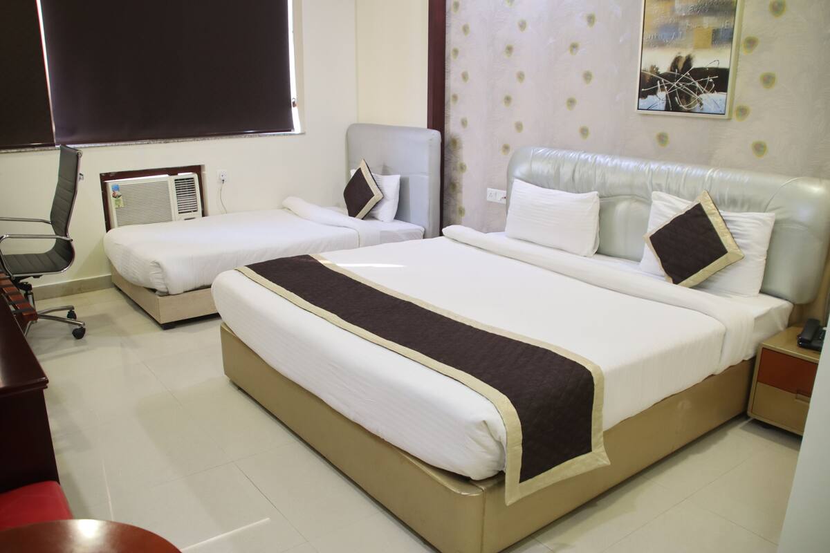 A hotel room with two single beds, a double bed, a desk with a chair, a nightstand, and a wall-mounted air conditioner.