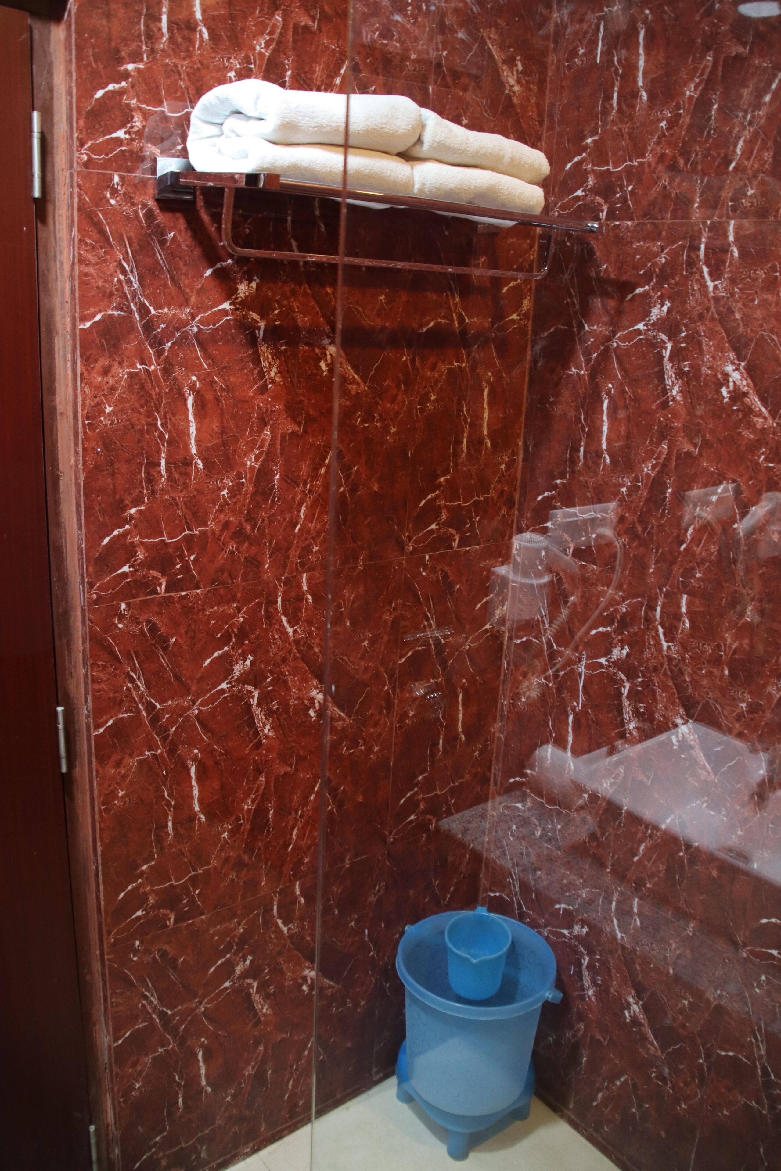 A bathroom with a marble wall, a towel rack with folded towels, and a blue bucket on the floor.
