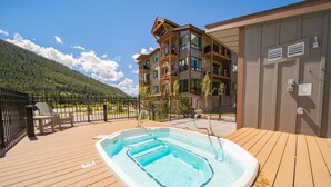 A wooden deck with a hot tub, a fenced area, and a multi-story building with balconies.