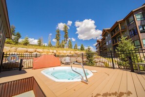Un jacuzzi en una terraza de madera con piscina, rodeado por una valla metálica, con montañas al fondo y un edificio con balcones.