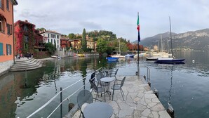 A lakeside terrace with stone flooring, metal chairs, and tables, overlooking a calm lake with boats and a flagpole. - Hotel La Pergola  (Bellagio)