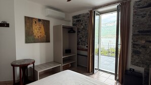 A bedroom with a bed, a ceiling fan, a painting, a wardrobe, a desk, a chair, a window with curtains, and a view of the outdoors. - Hotel La Pergola  (Bellagio)