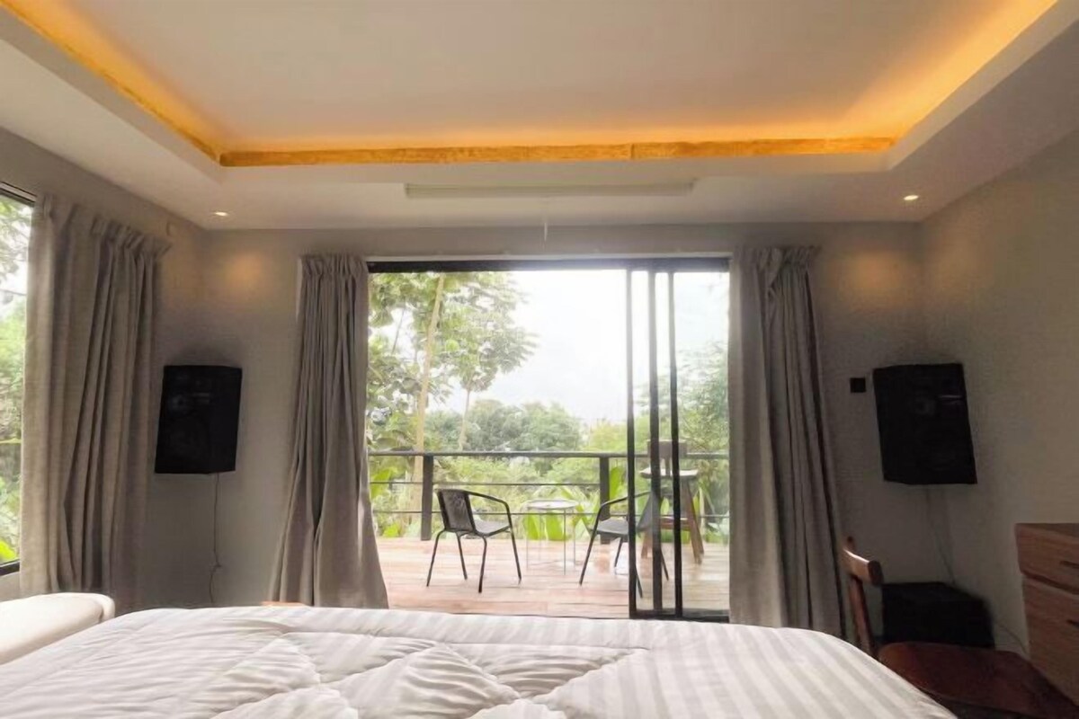 A hotel room with a large bed, a balcony with chairs, and a view of greenery.