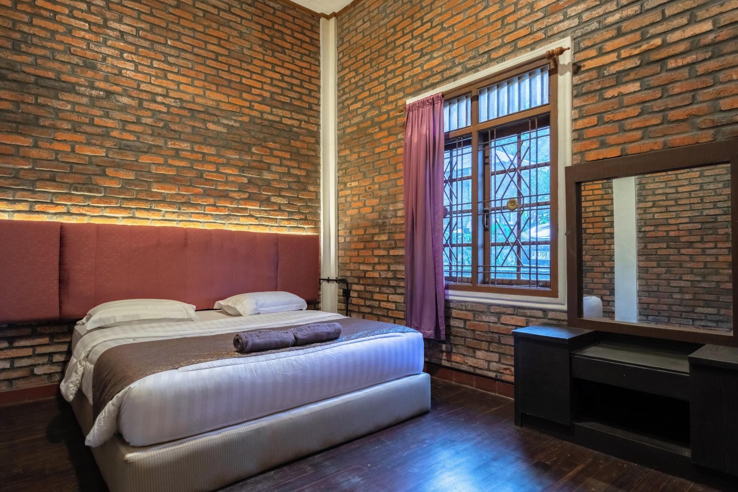 Una habitación con pared de ladrillo, una cama con ropa de cama blanca, una ventana con cortinas moradas y un aparador de madera oscura.