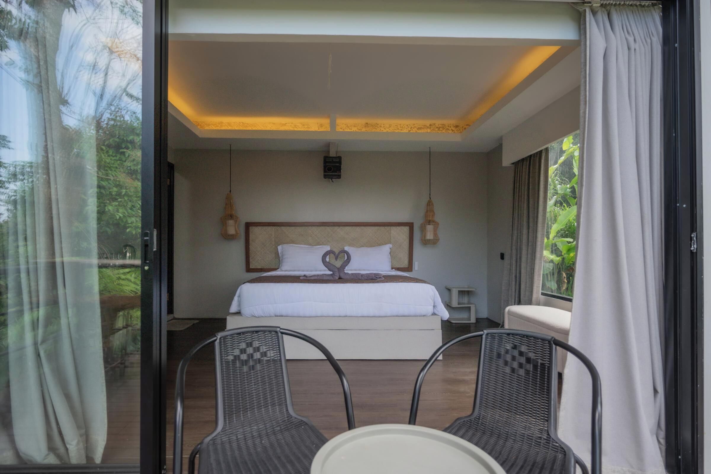 A bedroom with a bed, two bedside tables, a chandelier, and a view of greenery through a glass door.