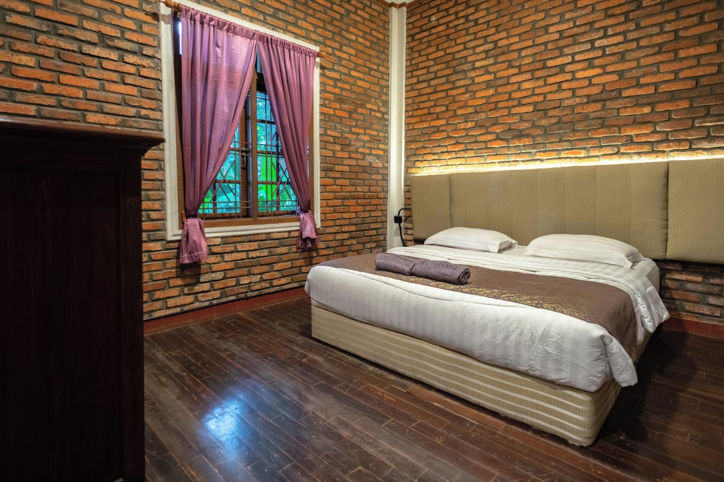 Un dormitorio con pared de ladrillo, una ventana con cortinas moradas, una cama con ropa de cama blanca y suelo de madera.