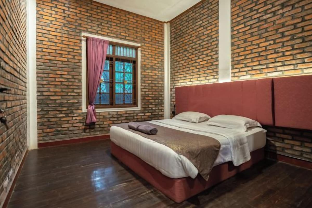 A bedroom with a brick wall, a window with curtains, a bed with pillows, and a wooden floor.