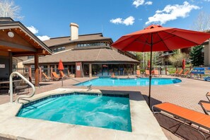 A pool area with lounge chairs and umbrellas, a hot tub, and a building with a sloped roof.