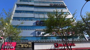 Un hotel de varios pisos con fachada de vidrio moderno, un letrero de restaurante a la vista y un comercio con toldo rojo.