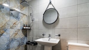 Un baño con pared de mármol, lavamanos, inodoro, cabezal de ducha de mano y espejo redondo.