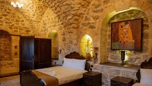 A stone-walled bedroom with a large bed, wooden furniture, and a painting on the wall. - Marinut Konağı (Mardin)