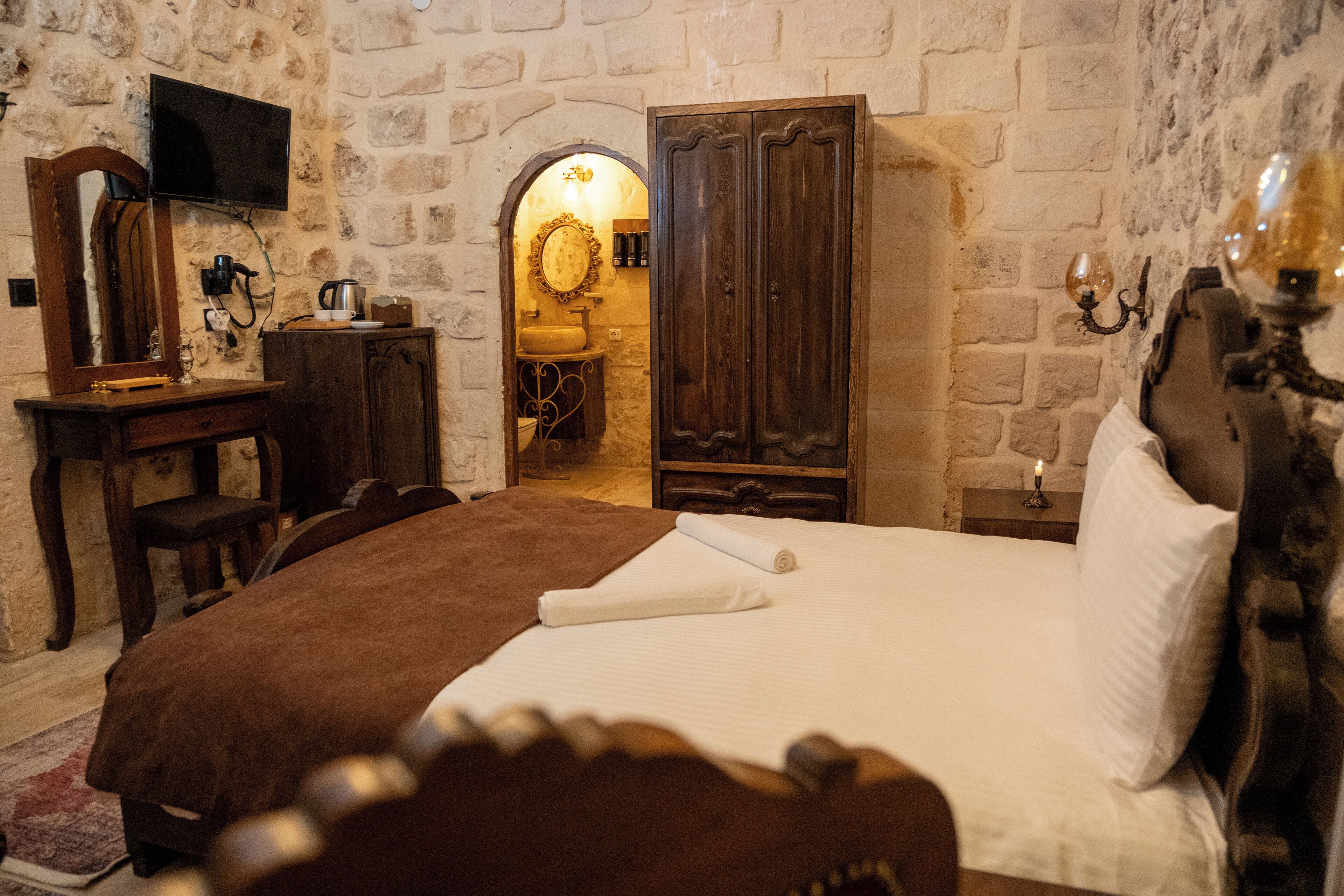 A bedroom with a large bed, a wooden dresser, a television mounted on the wall, and a stone wall.