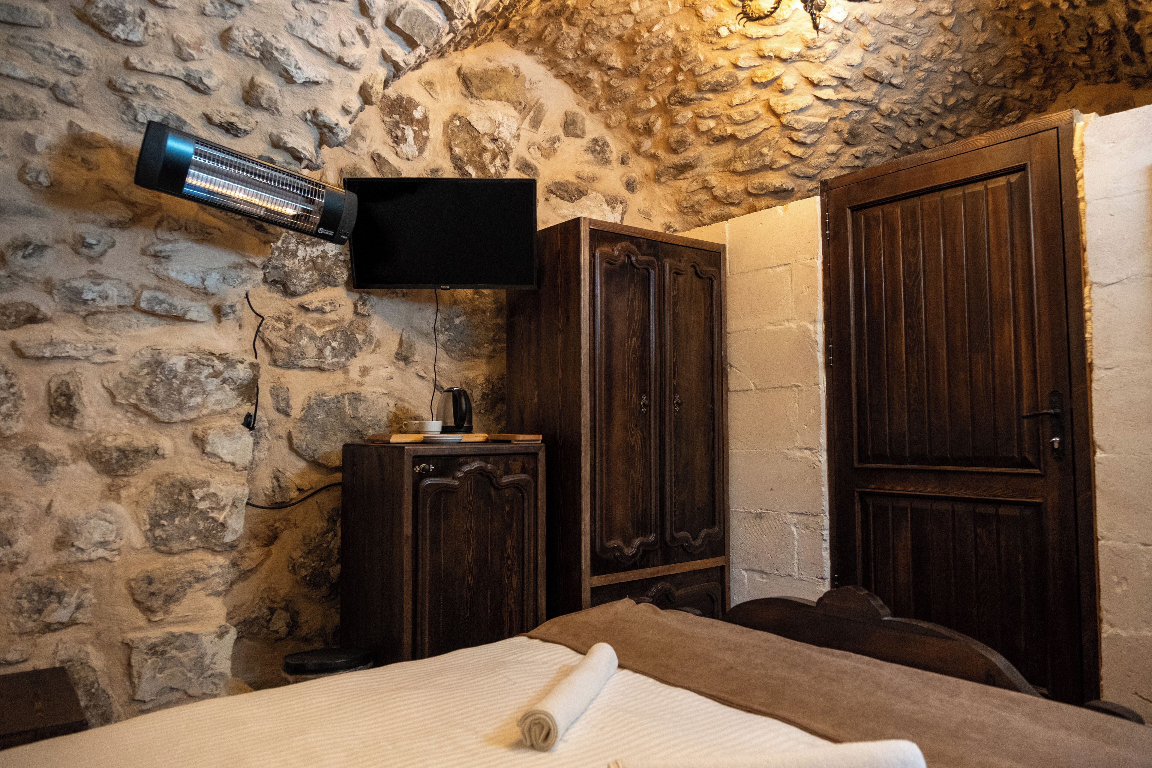 A stone-walled room with a bed, a television mounted on the wall, and wooden cabinets.