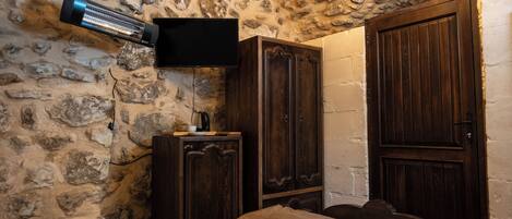 A stone-walled room with a bed, a television mounted on the wall, and wooden cabinets.