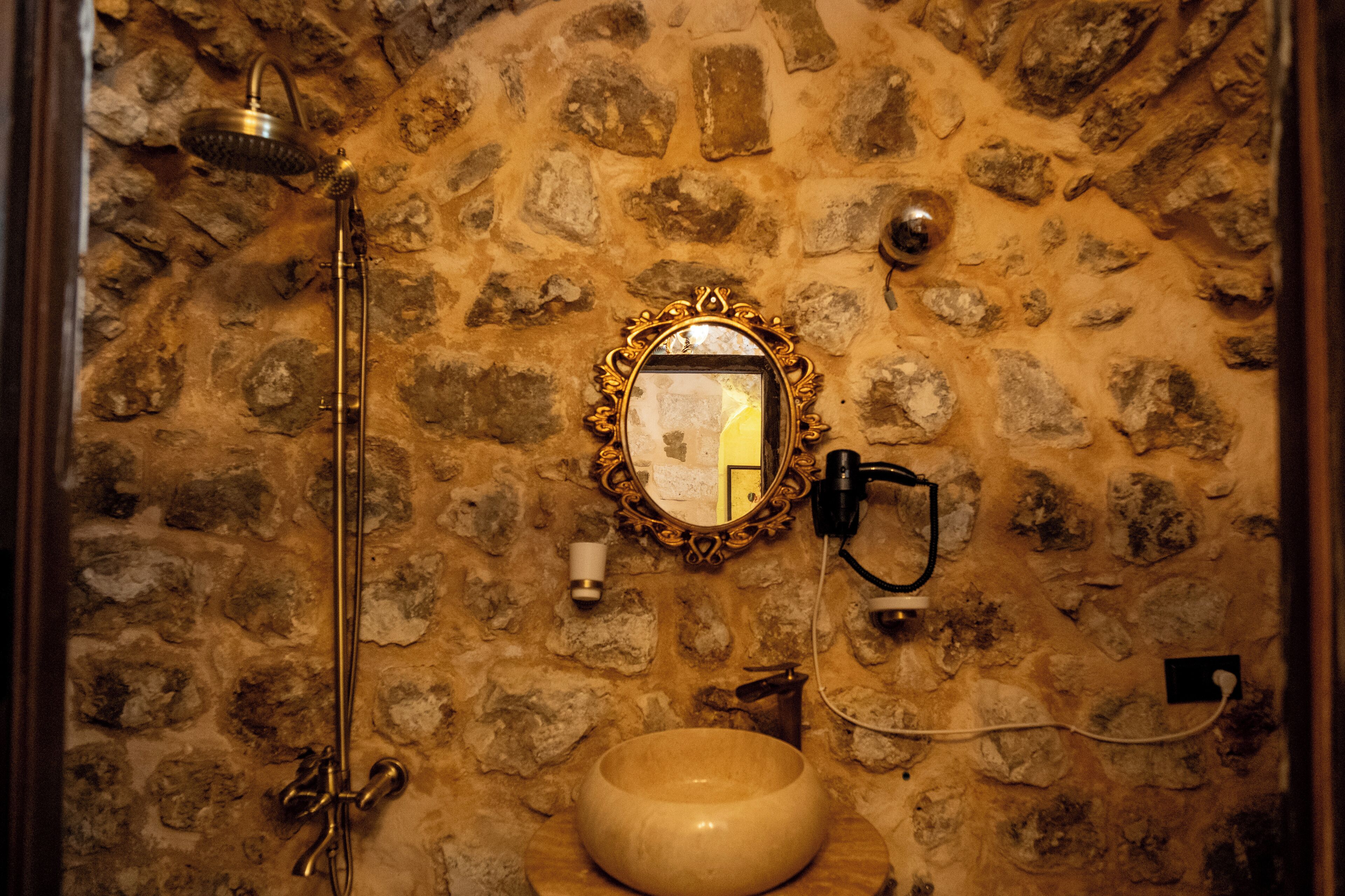 Un baño con paredes de piedra, un lavamanos de madera, un cabezal de ducha manual y un espejo instalado en la pared.